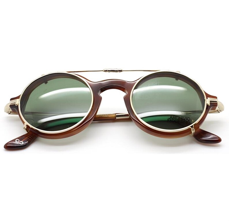 May include: Round sunglasses with a brown tortoiseshell frame and green lenses. The sunglasses have a gold-colored metal bridge and accents. The lenses have the text "ARCHIVO MUSEO" printed on them. The sunglasses are open and ready to wear.