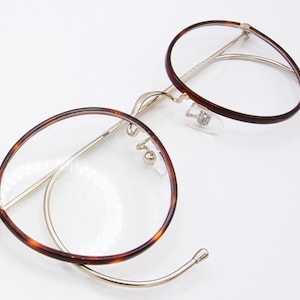 May include: Round eyeglasses with a tortoiseshell pattern on the rims and temples. The frames are a combination of gold-colored metal and the tortoiseshell design. The lenses are clear, and the glasses have nose pads.