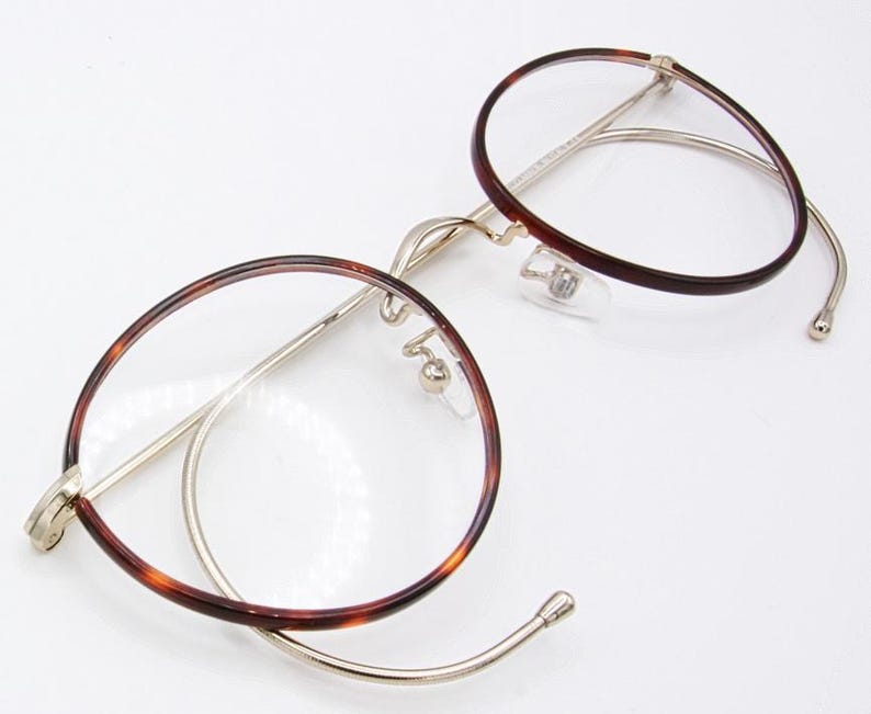 May include: Round eyeglasses with a tortoiseshell pattern on the rims and temples. The frames are a combination of gold-colored metal and the tortoiseshell design. The lenses are clear, and the glasses have nose pads.