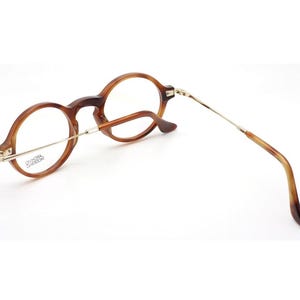 May include: Round-framed eyeglasses with a tortoiseshell pattern. The glasses have a gold-colored metal bridge and temples. The lenses are clear. The frame is a warm brown color. The glasses are angled, showing the side view.