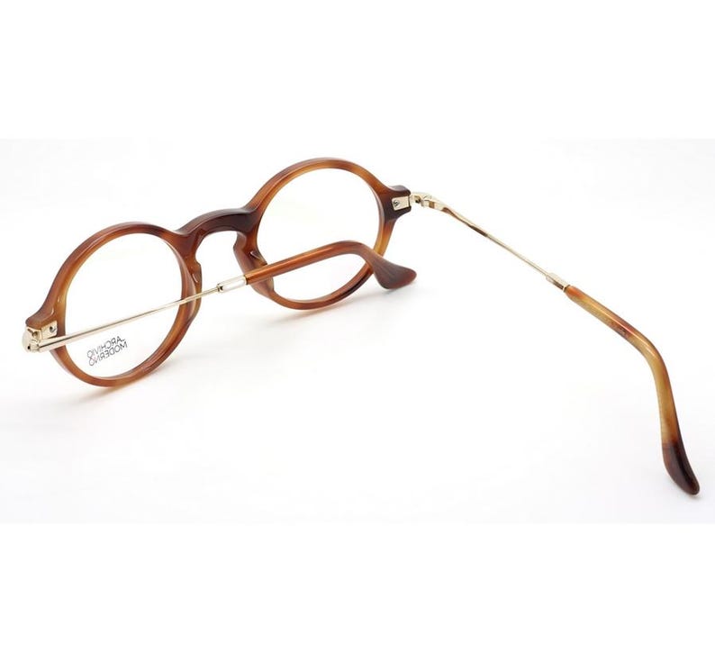 May include: Round-framed eyeglasses with a tortoiseshell pattern. The glasses have a gold-colored metal bridge and temples. The lenses are clear. The frame is a warm brown color. The glasses are angled, showing the side view.
