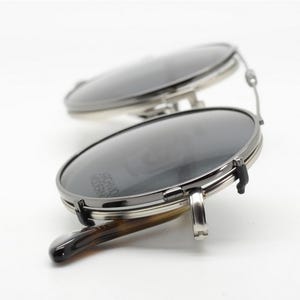 Puede incluir: Primer plano de unas gafas de sol redondas con lentes oscuras y montura plateada. Las patillas tienen un dise&ntilde;o de carey. El texto "ARCHIVO MODERNO" es visible.