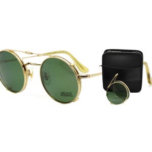 May include: Gold-rimmed sunglasses with green lenses and a matching case. The sunglasses have a double bridge and round lenses. The case is black and rectangular. The text "ARCHIVO MODERNO" is visible on the lens.