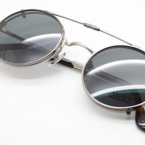 Puede incluir: Gafas de sol con montura plateada y lentes redondas y oscuras. Las gafas tienen un puente que conecta las dos lentes y patillas con estampado de carey. El texto "ARCHIVO MODERNO" es visible en la lente.