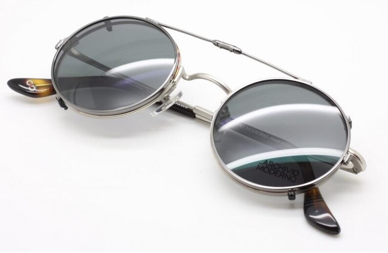 Puede incluir: Gafas de sol con montura plateada y lentes redondas y oscuras. Las gafas tienen un puente que conecta las dos lentes y patillas con estampado de carey. El texto "ARCHIVO MODERNO" es visible en la lente.