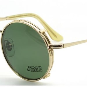 May include: Gold-rimmed sunglasses with green lenses. The round frames feature a double bridge and the text "ARCHIVO MODERNO" on the lower right lens. The temples and ear pieces are a light yellow color.