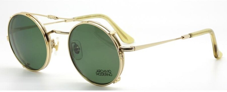 May include: Gold-rimmed sunglasses with green lenses. The round frames feature a double bridge and the text "ARCHIVO MODERNO" on the lower right lens. The temples and ear pieces are a light yellow color.