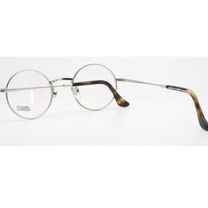 Puede incluir: Gafas con montura plateada, lentes redondas y dise&ntilde;o cl&aacute;sico. Las patillas presentan un patr&oacute;n de carey, a&ntilde;adiendo un toque vintage. Las gafas se presentan sobre un fondo blanco, resaltando su est&eacute;tica elegante y moderna.