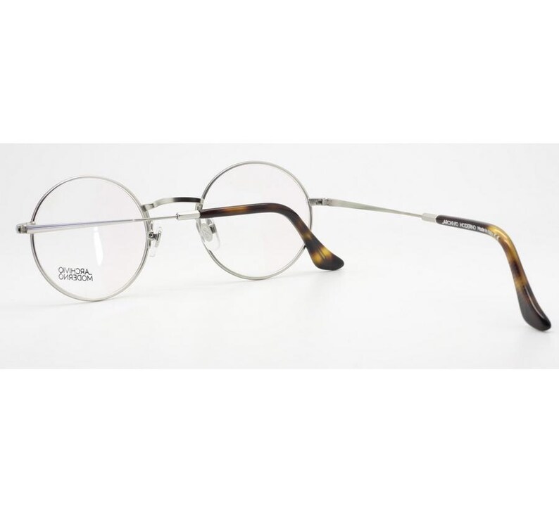 Puede incluir: Gafas con montura plateada, lentes redondas y dise&ntilde;o cl&aacute;sico. Las patillas presentan un patr&oacute;n de carey, a&ntilde;adiendo un toque vintage. Las gafas se presentan sobre un fondo blanco, resaltando su est&eacute;tica elegante y moderna.