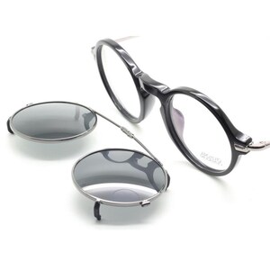 May include: Black round-frame eyeglasses with a clip-on sunglasses attachment. The eyeglasses have clear lenses and a dark frame. The clip-on sunglasses are oval-shaped with a dark tint and a silver frame.