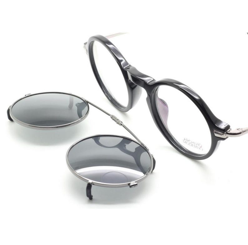May include: Black round-frame eyeglasses with a clip-on sunglasses attachment. The eyeglasses have clear lenses and a dark frame. The clip-on sunglasses are oval-shaped with a dark tint and a silver frame.
