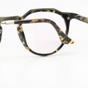 Puede incluir: Gafas con montura y patillas con estampado de carey. Las lentes son transparentes y las monturas tienen un dise&ntilde;o angular &uacute;nico. Las patillas est&aacute;n conectadas a las monturas con bisagras plateadas. El texto "DIVHORA MODERN" est&aacute; impreso en la lente.