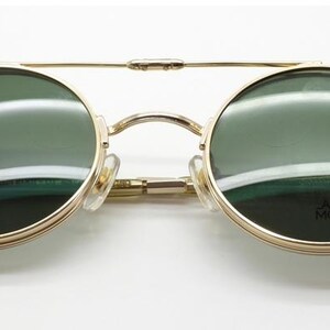 May include: Gold-framed sunglasses with round, green-tinted lenses. The glasses have a double bridge and light-colored earpieces. The text "ARCHIVE MODERN" is visible on the right lens.