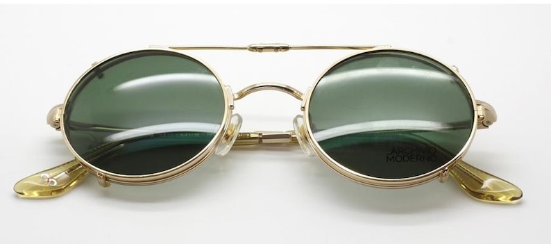 May include: Gold-framed sunglasses with round, green-tinted lenses. The glasses have a double bridge and light-colored earpieces. The text "ARCHIVE MODERN" is visible on the right lens.