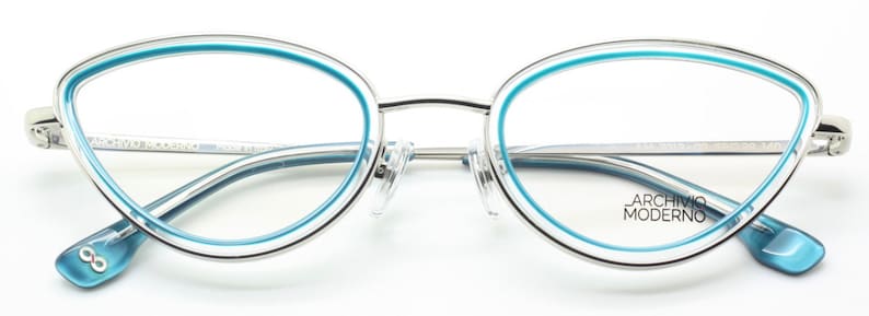 May include: Silver cat-eye glasses with a light blue trim. The glasses have a clear lens and the brand name "Archivio Moderno" is printed on the right temple.