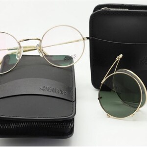 May include: A collection of eyewear, including round eyeglasses and folding sunglasses, with gold-colored frames. The glasses rest on black leather cases, one zippered and one with a flap. The brand name "ARCHIVO MODERNO" is visible on the cases.