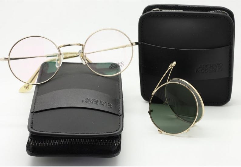 May include: A collection of eyewear, including round eyeglasses and folding sunglasses, with gold-colored frames. The glasses rest on black leather cases, one zippered and one with a flap. The brand name "ARCHIVO MODERNO" is visible on the cases.