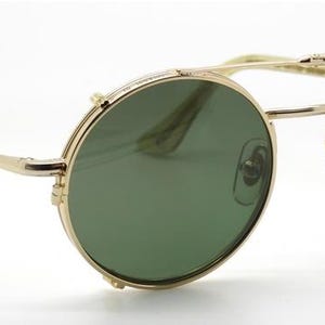 May include: Gold-rimmed sunglasses with round green lenses. The glasses have a double bridge and a classic, vintage design. The temples are thin and extend to the ear pieces. The lenses have a slight tint and the text "ARCHIVO MODERNO" is visible on the right lens.