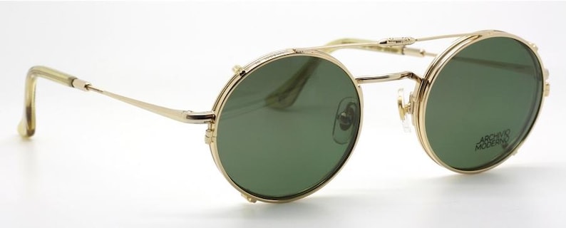 May include: Gold-rimmed sunglasses with round green lenses. The glasses have a double bridge and a classic, vintage design. The temples are thin and extend to the ear pieces. The lenses have a slight tint and the text "ARCHIVO MODERNO" is visible on the right lens.