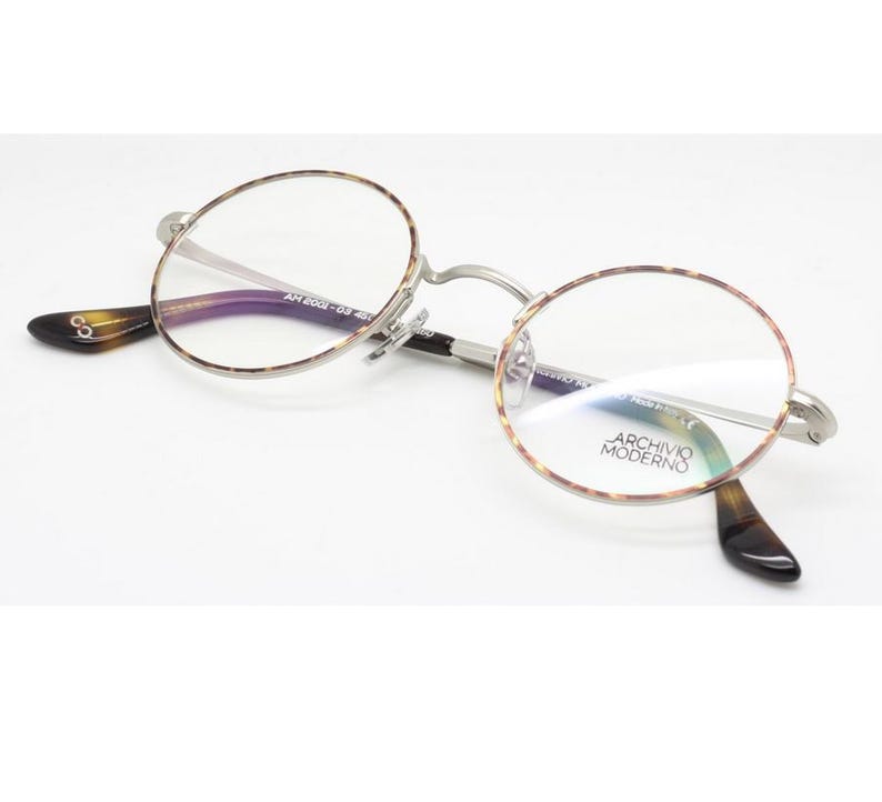 Puede incluir: Gafas redondas con montura plateada y detalles carey. Las lentes son transparentes y las patillas tienen un dise&ntilde;o marr&oacute;n oscuro y negro. La marca "ARCHIVIO MODERNO" es visible en la lente.