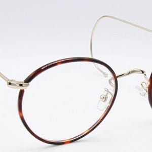 May include: Oval-shaped eyeglasses with a tortoiseshell pattern on the rims and a silver-colored metal frame. The glasses have a unique design with a curved temple arm that wraps around the ear. The lenses are clear.