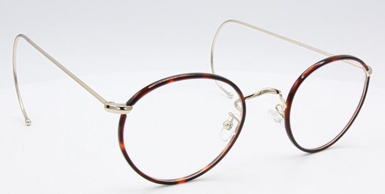 May include: Oval-shaped eyeglasses with a tortoiseshell pattern on the rims and a silver-colored metal frame. The glasses have a unique design with a curved temple arm that wraps around the ear. The lenses are clear.