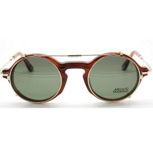 May include: Round brown sunglasses with gold-colored rims and a bridge. The lenses are green and the text "ARCHIVIO MODERNO" is printed on the right lens. The sunglasses have a vintage, retro style.