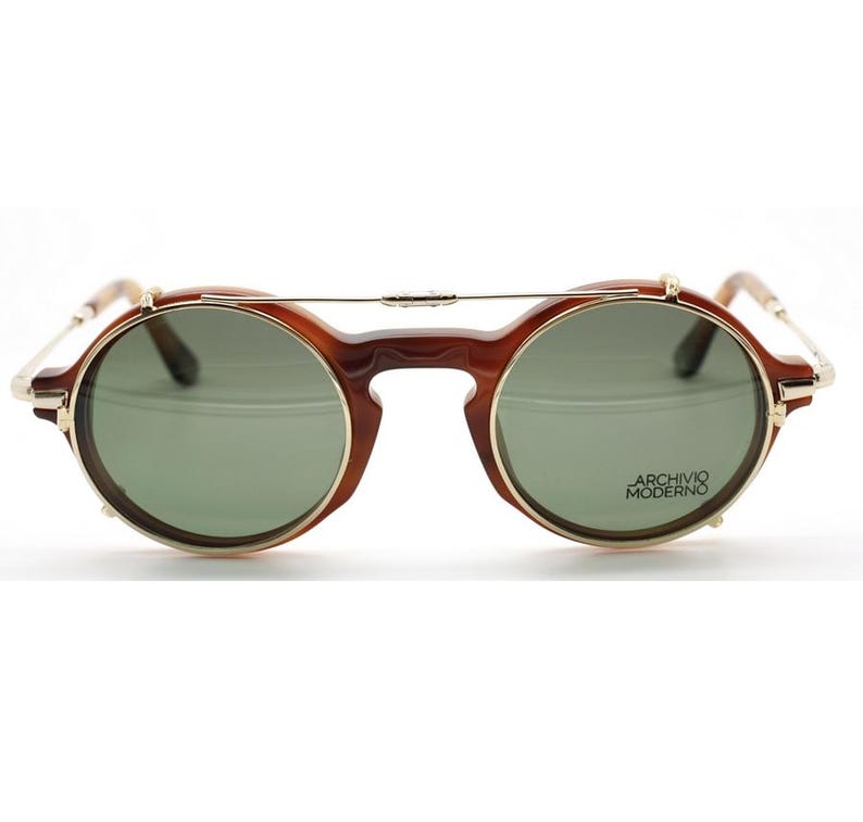 May include: Round brown sunglasses with gold-colored rims and a bridge. The lenses are green and the text "ARCHIVIO MODERNO" is printed on the right lens. The sunglasses have a vintage, retro style.