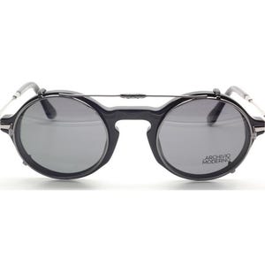 May include: Black round sunglasses with gray lenses and a silver bridge. The sunglasses have the text "ARCHIVO MODERNO" on the right lens. The frames are black with silver accents on the temples.