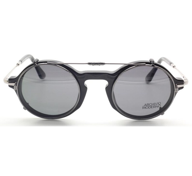 May include: Black round sunglasses with gray lenses and a silver bridge. The sunglasses have the text "ARCHIVO MODERNO" on the right lens. The frames are black with silver accents on the temples.