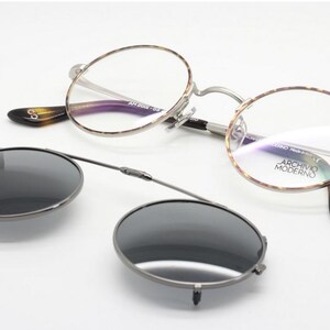 Puede incluir: Gafas redondas con un accesorio de gafas de sol con clip. Las gafas tienen una montura plateada y patillas con estampado de carey. Las gafas de sol con clip son de color gris oscuro.