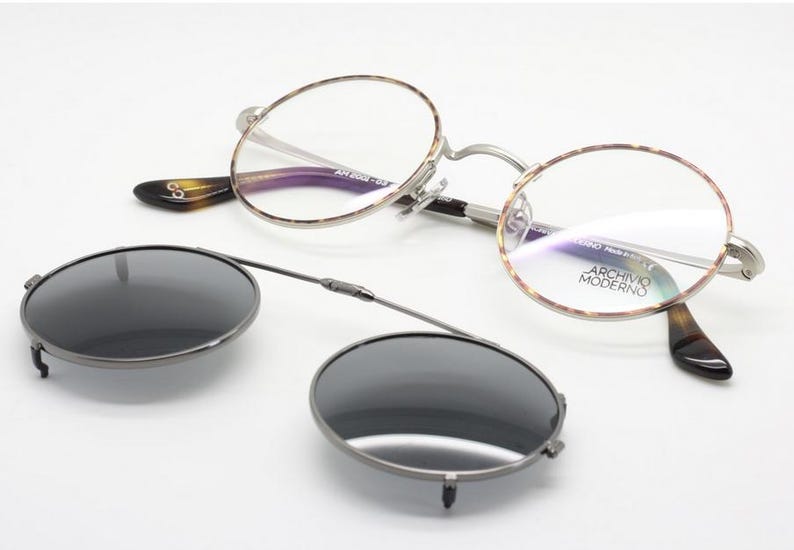 Puede incluir: Gafas redondas con un accesorio de gafas de sol con clip. Las gafas tienen una montura plateada y patillas con estampado de carey. Las gafas de sol con clip son de color gris oscuro.