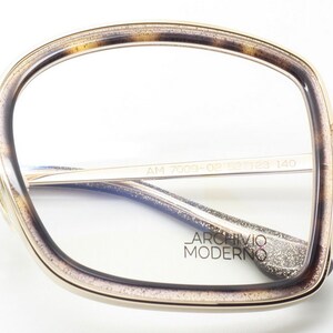 May include: Gold-rimmed eyeglasses with a tortoise shell pattern on the frame. The glasses have a clear lens and the text "AM 7009-02 53 18 140 ARCHIVIO MODERNO" is printed on the inside of the frame.