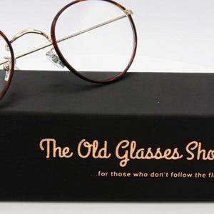 May include: Round eyeglasses with a tortoiseshell pattern on the rims rest on a black box. The box has gold lettering that reads "The Old Glasses Shop" and the tagline "for those who don't follow the flock".