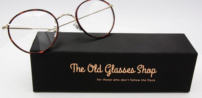 May include: Round eyeglasses with a tortoiseshell pattern on the rims rest on a black box. The box has gold lettering that reads "The Old Glasses Shop" and the tagline "for those who don't follow the flock".
