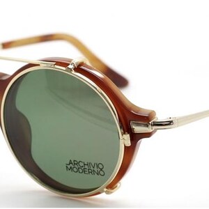 May include: Brown and gold-tone sunglasses with round green lenses. The sunglasses have a bridge and temples with a gold-tone metal frame. The text "ARCHIVO MODERNO" is printed on the lens.