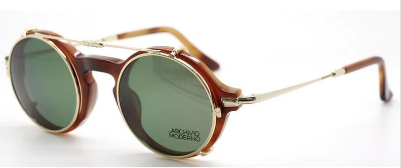 May include: Brown and gold-tone sunglasses with round green lenses. The sunglasses have a bridge and temples with a gold-tone metal frame. The text "ARCHIVO MODERNO" is printed on the lens.