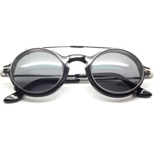May include: Round black sunglasses with a silver bridge and arms. The lenses are a dark gray color, and the frame has a modern design. The text "ARCHIVIO MODERNO" is visible on the arm.