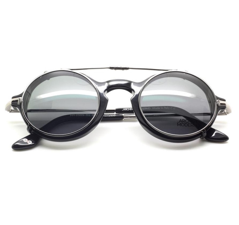 May include: Round black sunglasses with a silver bridge and arms. The lenses are a dark gray color, and the frame has a modern design. The text "ARCHIVIO MODERNO" is visible on the arm.