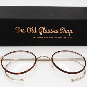 May include: Round eyeglasses with a gold-colored metal frame and tortoise-shell patterned rims. The glasses are displayed in front of a black box with the text "The Old Glasses Shop" and the tagline "for those who don't follow the flock".