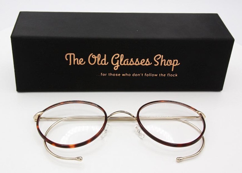May include: Round eyeglasses with a gold-colored metal frame and tortoise-shell patterned rims. The glasses are displayed in front of a black box with the text "The Old Glasses Shop" and the tagline "for those who don't follow the flock".