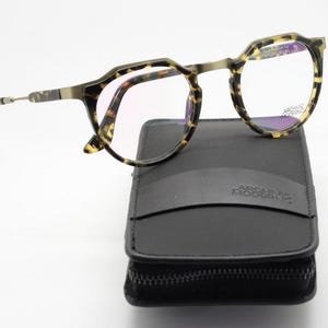 Puede incluir: Gafas con montura con estampado de carey sobre un estuche negro con cremallera. Las gafas tienen una forma geom&eacute;trica &uacute;nica y lentes transparentes. El estuche lleva la inscripci&oacute;n "ARCHIVO MODERNO".