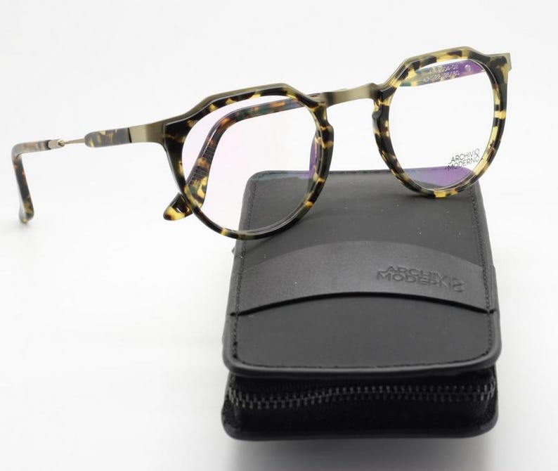Puede incluir: Gafas con montura con estampado de carey sobre un estuche negro con cremallera. Las gafas tienen una forma geom&eacute;trica &uacute;nica y lentes transparentes. El estuche lleva la inscripci&oacute;n "ARCHIVO MODERNO".