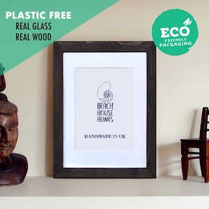 May include: A dark grey wood picture frame with a white mount and a white background. The frame is on a shelf with a wooden statue and a small wooden chair. The frame has the text "Beach House Frames" and "Handmade in UK" printed on the white background.