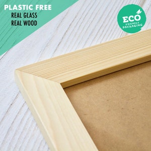 May include: Close-up of a natural wood picture frame with a brown cardboard backing. The frame is made of real wood and glass. The text "Plastic Free, Real Glass, Real Wood" is visible in the top left corner of the image. The text "Eco Friendly Packaging" is visible in the top right corner of the image.