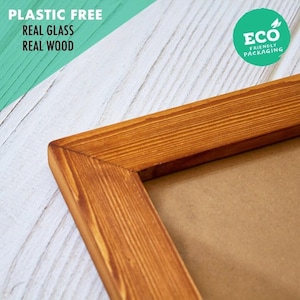 May include: A rectangular picture frame made of natural brown wood with a visible grain pattern. The frame features a mitered corner joint and a smooth finish. Text on the image reads: PLASTIC FREE, REAL GLASS, REAL WOOD, and ECO PACKAGING.