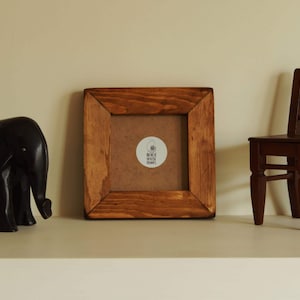 May include: A square wooden picture frame with a dark brown stained finish and visible wood grain. The centre of the frame contains a circular white sticker with a black nautilus shell icon and the text BEACH HOUSE FRAMES printed in black.