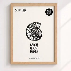 The image is a framed artwork featuring a black and white illustration of a spiral shell or nautilus shell. The text on the frame reads "SOLID OAK" and "BEACH HOUSE FRAMES" along with "HANDMADE IN THE UK".