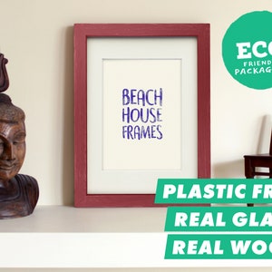 May include: A red wood picture frame with a white mount and a white background. The text "Beach House Frames" is printed in purple on the white background. The frame is sitting on a white shelf with a wooden statue and a small wooden chair. The text "Plastic Free, Real Glass, Real Wood" is printed in green on a white background below the frame.