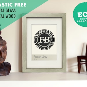 Op de afbeelding: Een lichtgrijze houten fotolijst met een witte passe-partout en een zwart-wit logo voor Farrow & Ball Paint & Paper. Het logo zegt "French Gray" en "No. 18". De lijst staat op een witte plank met een houten beeld en een kleine houten stoel.